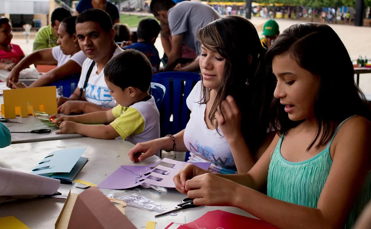Dos jóvenes, un niño, una mujer y un hombre reunidos están en una mesa, participando de una actividad con cartulinas y tijeras.